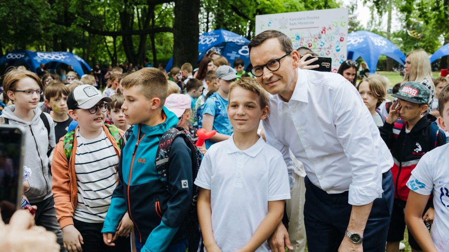 Mateusz Morawiecki, wówczas premier, pozuje z dziećmi podczas wydarzenia programu 800+ na zielonym terenie