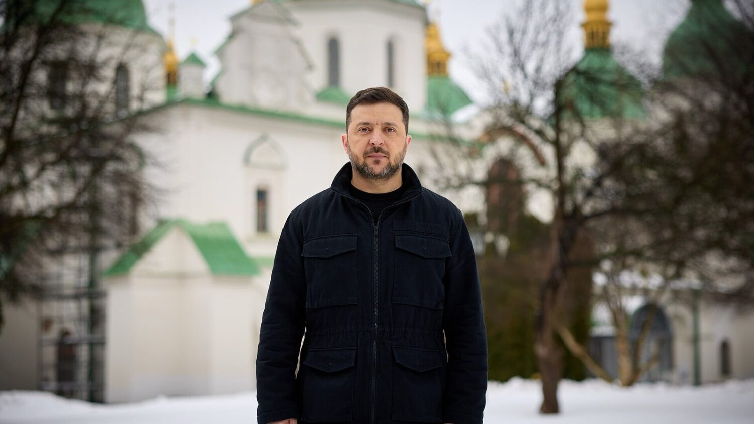 Wołodymyr Zełenski w czarnej kurtce stoi zimą na śniegu przed cerkwią ze złotymi kopułami w Kijowie