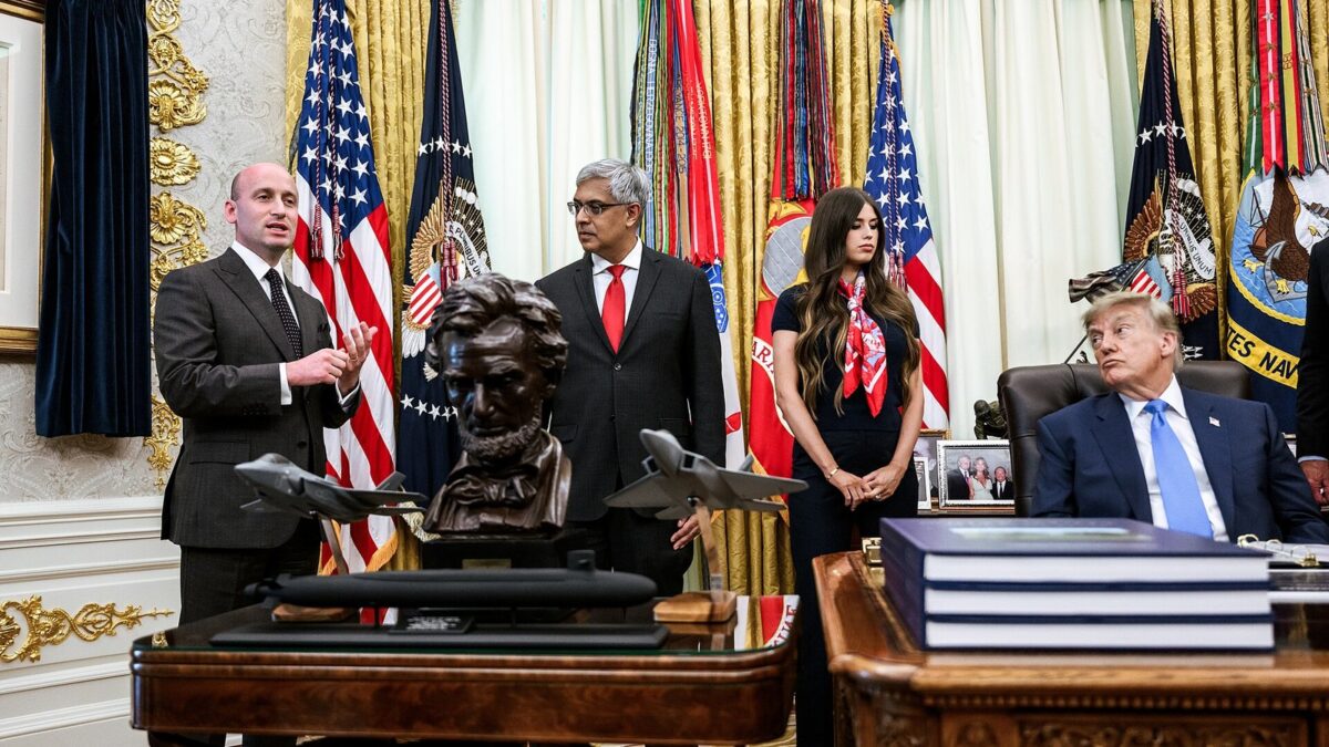 Donald Trump siedzi za biurkiem w Gabinecie Owalnym Białego Domu, obok stoją goście i mówca.
