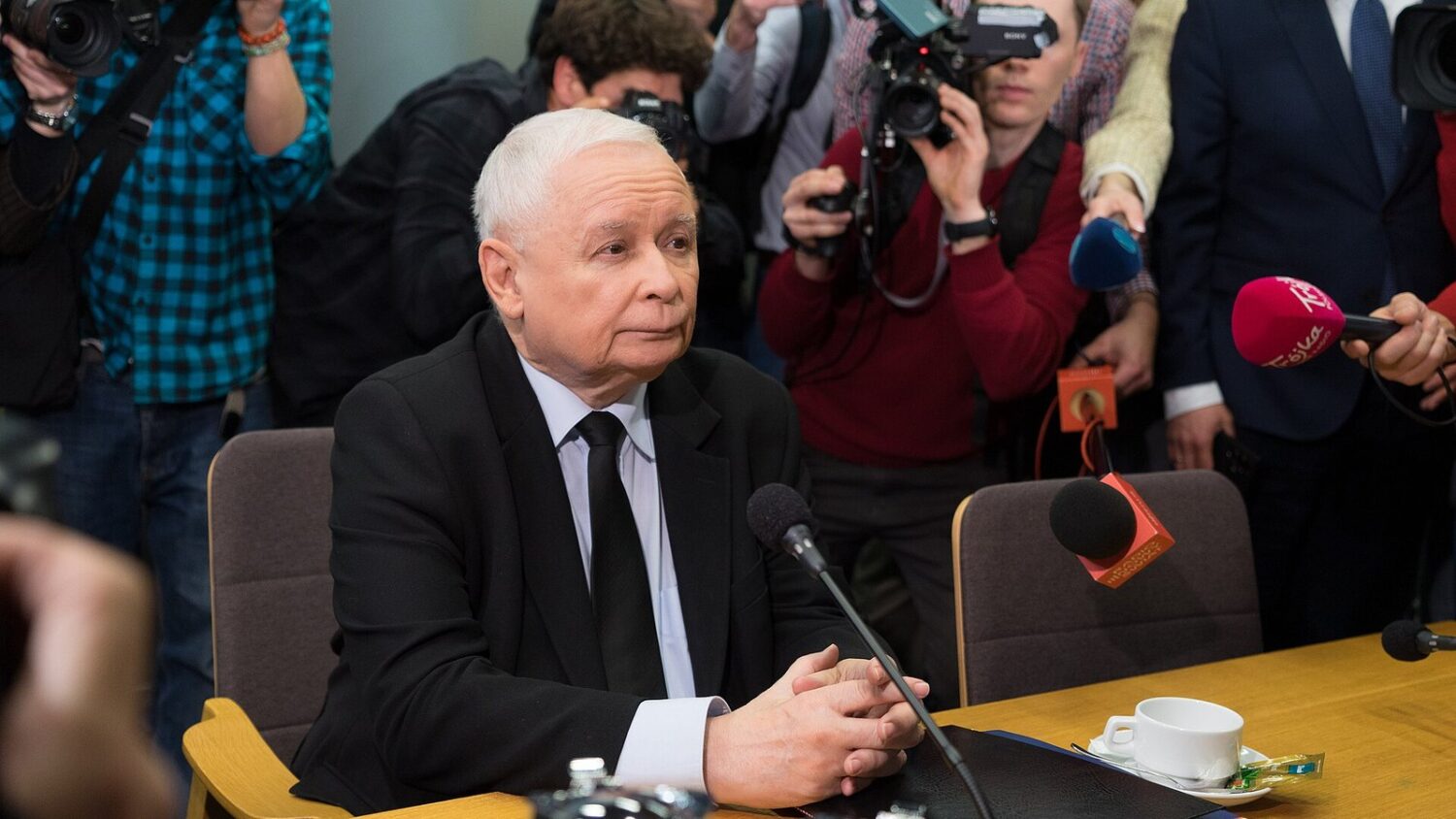 Jarosław Kaczyński przy stole z mikrofonami podczas spotkania z mediami; w tle dziennikarze z kamerami.