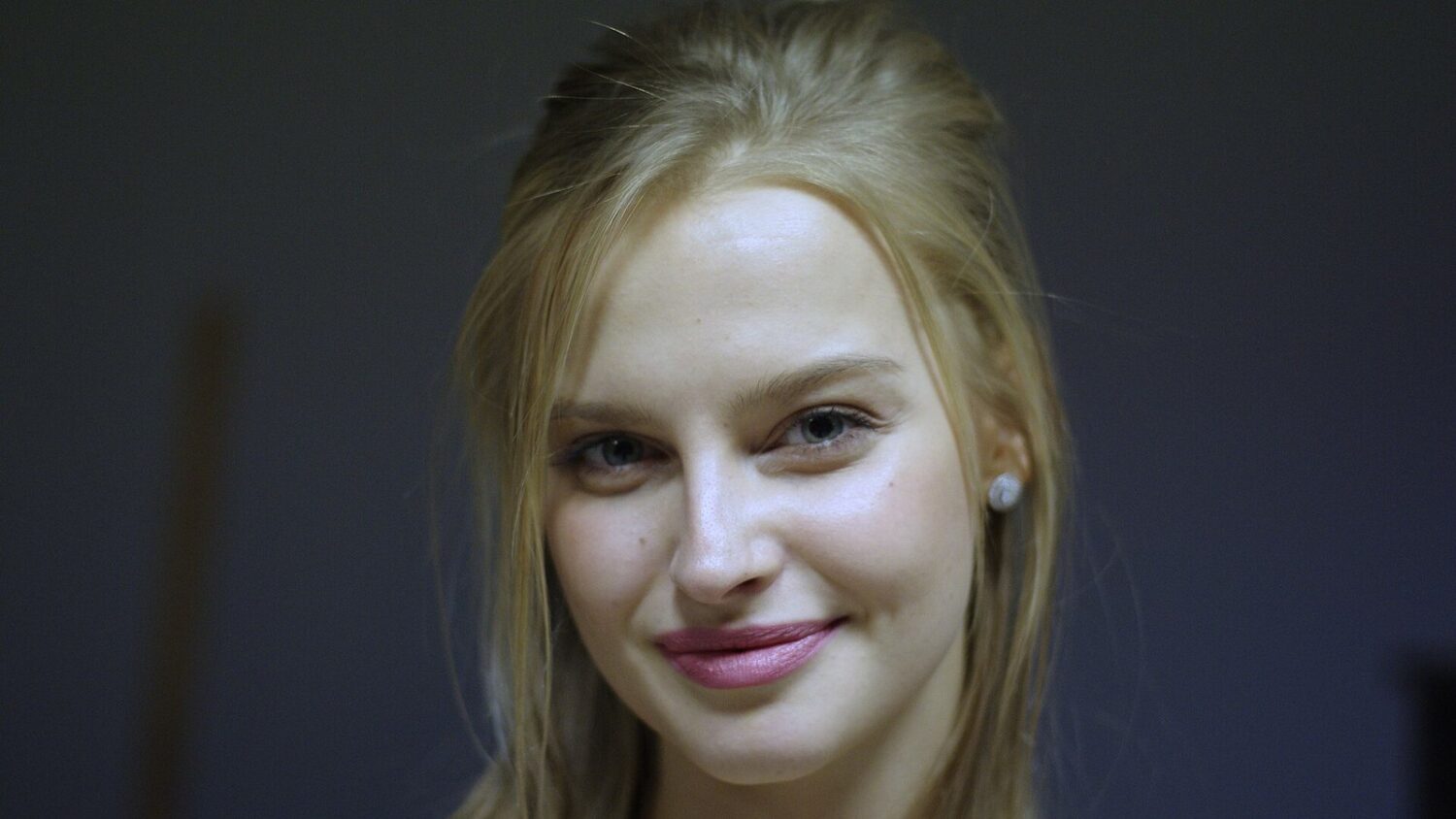 Karolina Pisarek, blondynka z rozpuszczonymi włosami i perłowym kolczykiem, uśmiecha się do kamery na ciemnym tle.