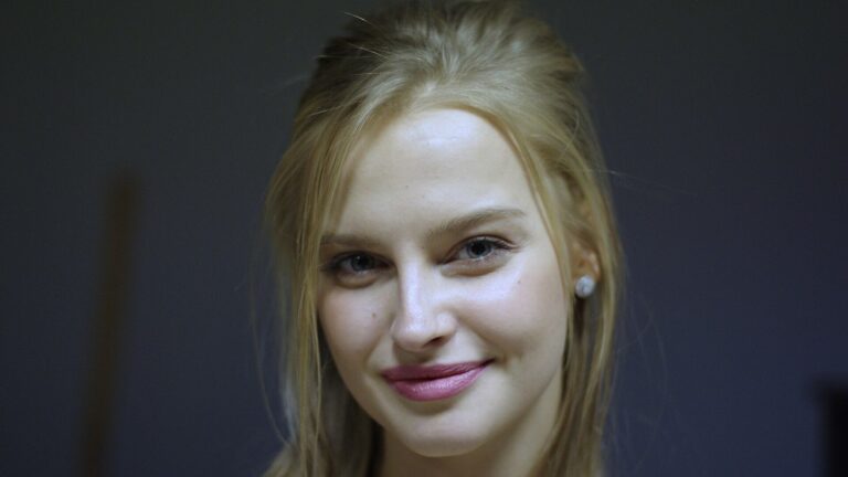 Karolina Pisarek, blondynka z rozpuszczonymi włosami i perłowym kolczykiem, uśmiecha się do kamery na ciemnym tle.