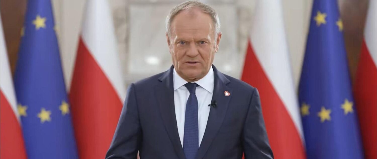 Premier Donald Tusk w granatowym garniturze przemawia na tle flag Polski i Unii Europejskiej