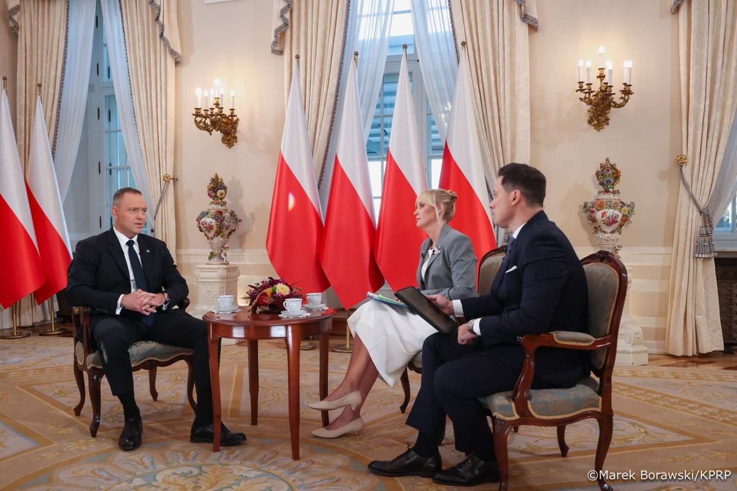 Prezydent Karol Nawrocki podczas wywiadu w reprezentacyjnej sali; w tle biało-czerwone flagi.