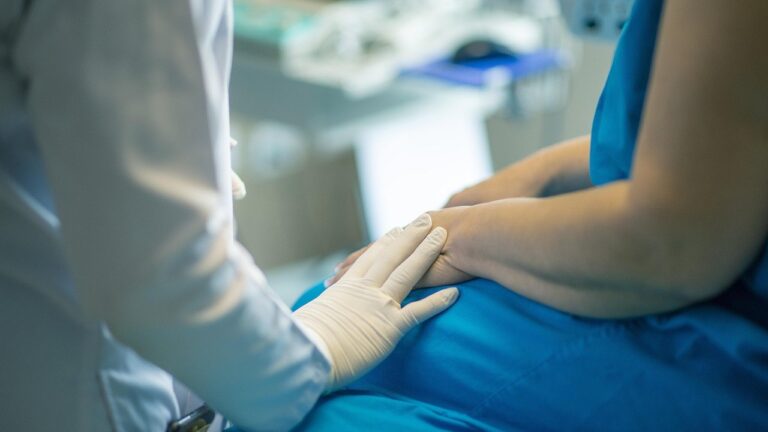 Lekarz w białym fartuchu i rękawiczkach trzyma dłoń pacjenta leżącego w niebieskiej pościeli.