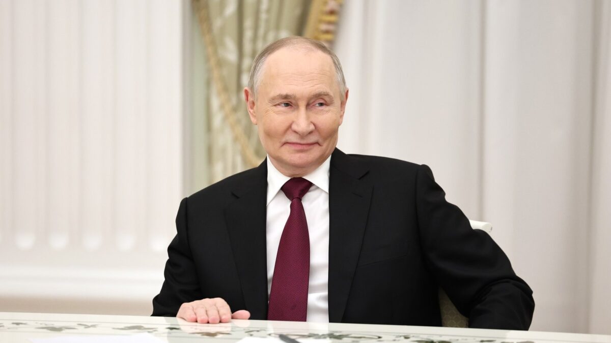 Władimir Putin siedzi przy stole na Kremlu i uśmiecha się podczas spotkania.