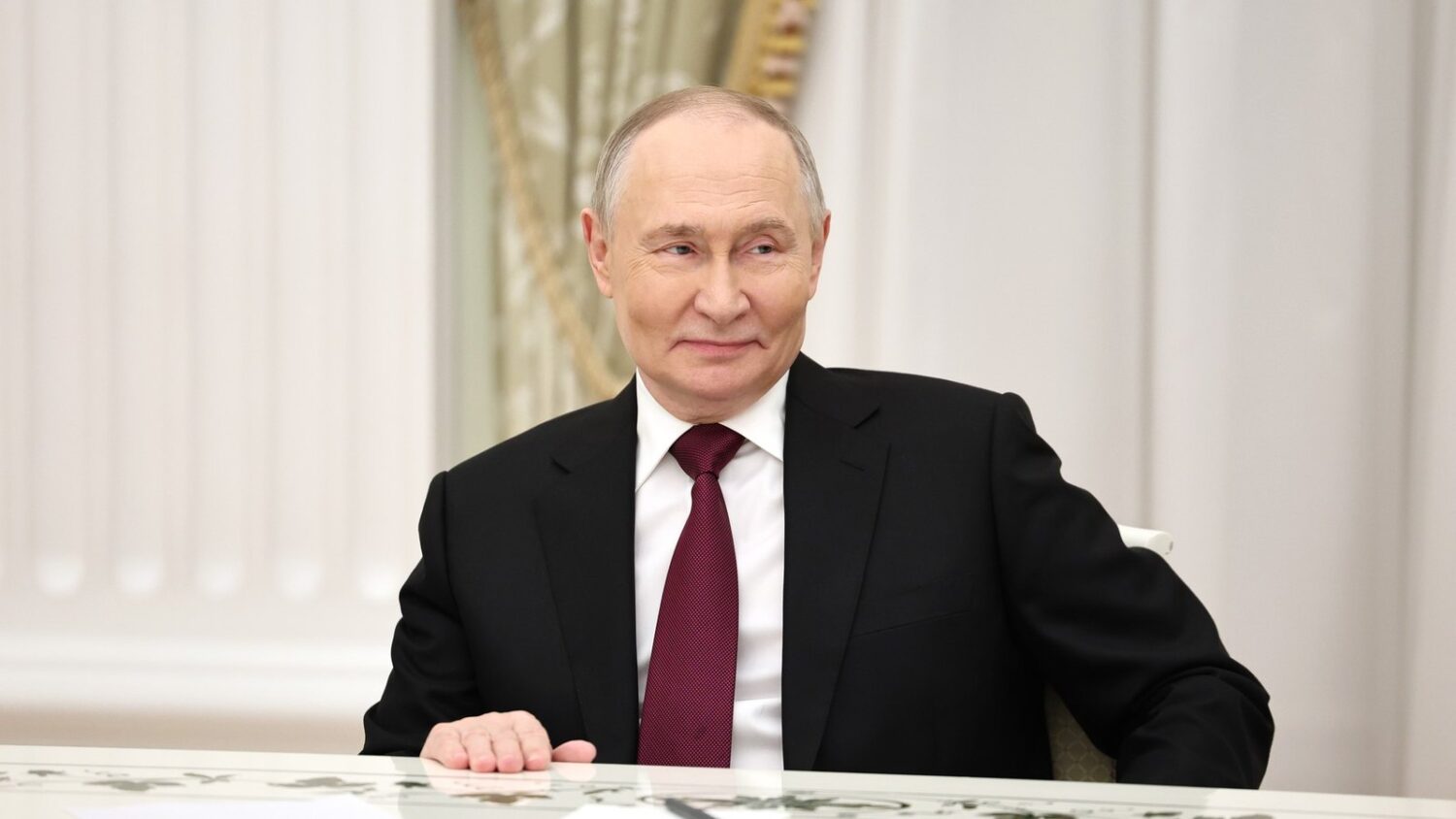 Władimir Putin siedzi przy stole na Kremlu i uśmiecha się podczas spotkania.